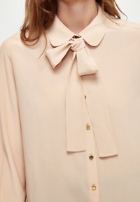 Blusa rosa pesca chiaro con colletto a fiocco, maniche lunghe e una fila di bottoni gialli lungo il davanti. Tessuto liscio.