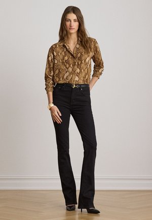 Kvinde, der står indendørs iført en brun bluse med slangeprint, sorte high-waisted jeans, sorte hæle og et gyldent armbånd.