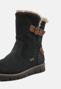 Bottine en suède noir avec doublure en fausse fourrure, accents en cuir marron, boucle décorative et semelle en caoutchouc texturée. Marque Gore-Tex visible.