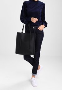 Kvinna i mörkblå långärmad topp och jeans som håller en stor svart tygväska, iförd vita sneakers på vit bakgrund.