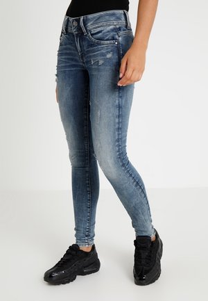 Persona que lleva vaqueros pitillo azules desgastados y zapatillas deportivas negras, de pie sobre un fondo claro y liso.