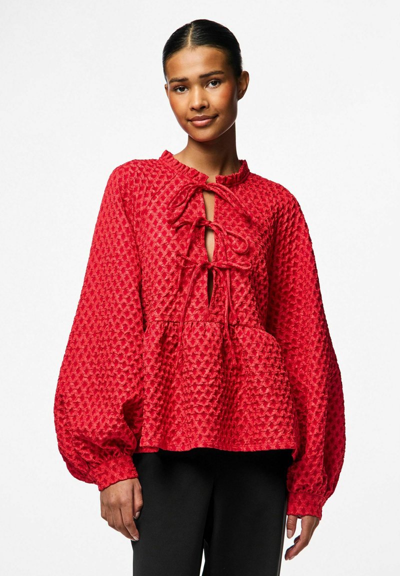 Punane tekstuuriga pluus koos volangkraega, võtmeaugu dekolteega ja nööriga kaunistustega. Omab mahukaid pikaid varrukaid ja peplum-siluetti.