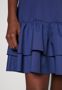 Mörkblå klänning med ett flerlagers volangnederdel, tillverkad av ett mjukt tyg. Volangerna är jämnt lager på lager med en samlad söm vid midjan.