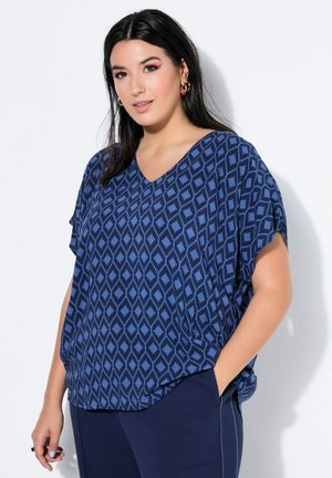 Kvinde med langt mørkt hår iført blåt mønstret kortærmet bluse og mørkeblå bukser, stående med den ene hånd i lommen mod hvid baggrund.