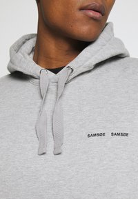 Hanorac gri cu cordoane reglabile, textură herringbone și logo-ul brodat negru "SAMØE SAMØE" pe piept. Material moale, design casual.