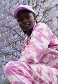Conjunto de jaqueta e calças em tie-dye rosa e branco, com um logo, usado com um boné a condizer. O fundo inclui uma árvore despida e um edifício.