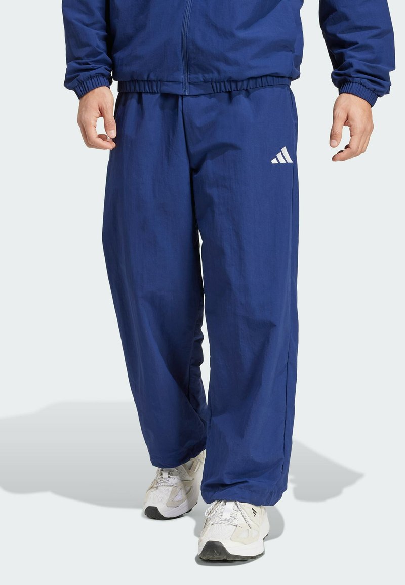 Marineblå letvægts træningsbukser med elastisk talje og åbninger, prydet med hvid Adidas-logo på fronten, parret med hvide sportssko.