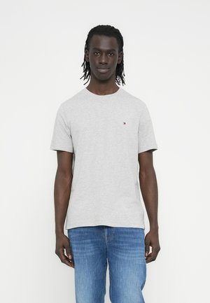 Mann mit mittel-langen Dreadlocks, der ein hellgraues Kurzarm-T-Shirt und blaue Jeans trägt, steht vor einem schlichten weißen Hintergrund.