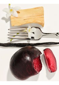 Fork with white flower, wooden block, vanilla pod, and a dark purple plum with a sliced piece showing red flesh on a white background.