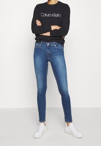 Tmavě modré skinny džíny doplněné černou mikinou s kulatým výstřihem s bílým logem "Calvin Klein", k tomu bílé tenisky.