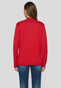 Roter Pullover mit langen Ärmeln, gerippter Struktur, rundem Halsausschnitt und lockerem Schnitt. Getragen mit blauen Denim-Jeans.