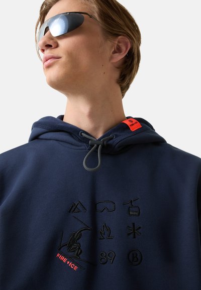 Mladý člověk nosící reflexní sluneční brýle a tmavě modrou mikinu s černými zimními ikonami a červeným textem "FIRE + ICE" na hrudi a kapuci.