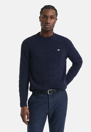 Marineblauer Kabelstrickpullover mit Rundhalsausschnitt und strukturiertem Muster. Verfügt über ein kleines Logo auf der Brust, kombiniert mit dunklen Hosen.