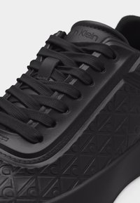 Μαύρο αθλητικό παπούτσι Calvin Klein με ανάγλυφο μοτίβο λογότυπου, μαύρα κορδόνια και λείο δερμάτινο γλώσσο που φέρει το όνομα της μάρκας.