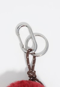 Czerwony futrzany breloczek z pomponem z brązową plecioną taśmą, srebrnym metalowym klipsem karabinkowym i detalem pierścienia na klucze. Miękka tekstura z bezpiecznym mocowaniem.