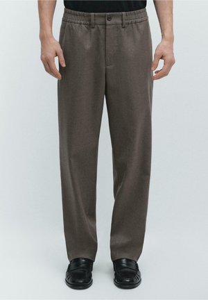 Mann trägt braune gerade geschnittene Hose mit Knopf und elastischem Bund, kombiniert mit schwarzen Schuhen, steht vor einfarbigem Hintergrund.