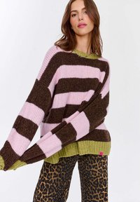 Gruby sweter z dzianiny w różowe i brązowe paski, z zielonym prążkowanym wykończeniem i długimi rękawami, w zestawieniu z spodniami w panterkę.