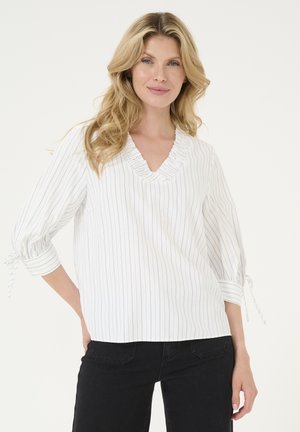 Weiße Bluse mit vertikalen blauen Streifen, V-Ausschnitt und Rüschensaum. Mit 3/4-Ärmeln und gebundenen Manschetten. Kombiniert mit schwarzen Hosen.