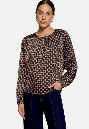 Femme portant un chemisier marron à manches longues avec des motifs circulaires blancs et un jean bleu foncé, debout avec une main dans la poche.