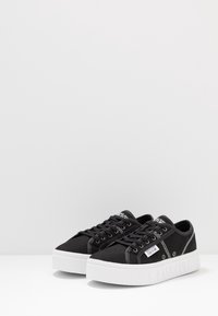 Černé nízké plátěné tenisky s bílými silnými podrážkami a značkovými štítky na boku a jazyku, umístěné na bílé ploše.