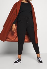 Pantaloni neri aderenti con vita elasticizzata, abbinati a un maglione nero a trama e a un lungo cappotto color ruggine con bottoni e fodera in raso. Scarpe da ginnastica nere completano l'outfit.