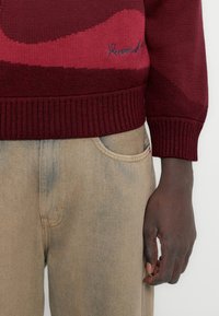 Μπορντό και ροζ πλεκτό πουλόβερ με ριγιέ μανσέτες, που διαθέτει κυματιστό μοτίβο και διακριτική σήμανση. Συνδυασμένο με ανοιχτό μπεζ τζιν.