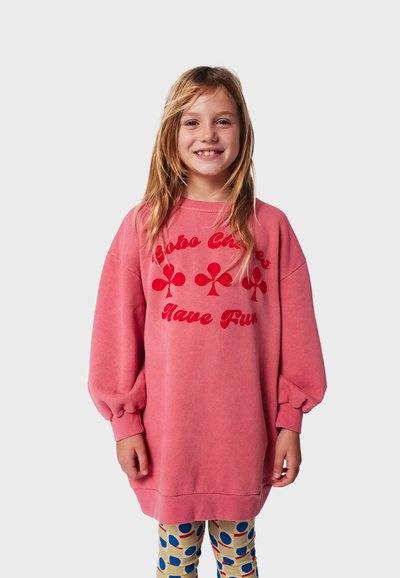 Felpa oversize rosa con testo stampato rosso e motivi di trifoglio, abbinata a leggings a fantasia con cerchi su una griglia.