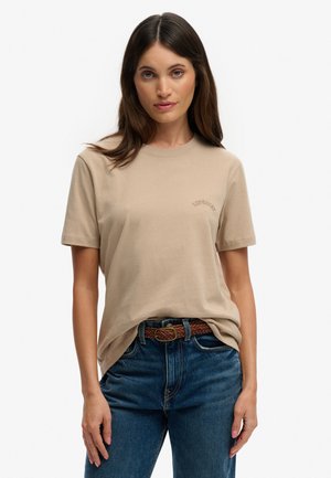 Kvinde med langt brunt hår iført en beige kortærmet t-shirt stoppet ned i blå jeans med et vævet brunt bælte mod en ensfarvet hvid baggrund.