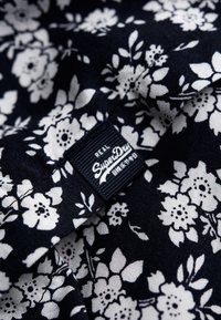 Tissu marine avec un motif floral blanc, présentant une étiquette rectangulaire sombre avec "SuperDry" en texte blanc et des détails fins.