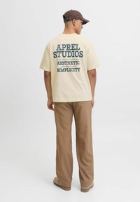 T-shirt crem cu imprimeu pe spate în text albastru închis, având inscripția "APREL STUDIOS" și diverse fraze. Croială lejeră, asortată cu pantaloni largi de culoare maro.