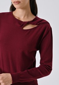 Donna che indossa un maglione bordeaux a maniche lunghe con un dettaglio intrecciato vicino al colletto, in piedi contro uno sfondo semplice.