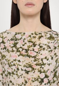 Femme portant un haut beige à motifs floraux avec des feuilles vertes et des fleurs roses, visible des épaules aux lèvres, avec des cheveux droits et foncés.