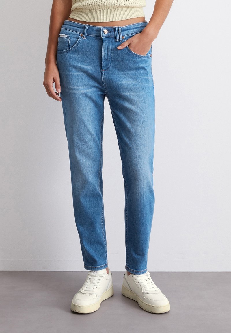 Jasnoniebieskie dżinsy o prostym kroju nogawki, pięć kieszeni i zapięcie na guzik, zestawione z białymi trampkami i jasnym swetrem.