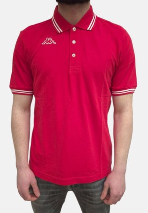 Homme portant un polo rouge à manches courtes avec col et poignets blancs à rayures, debout devant un fond uni.