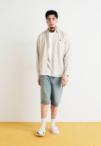 Lys beige skjorte med knapper over en hvit t-skjorte, paret med falmede blå denimshorts og hvite sokker med sandaler. Enkelt bakgrunn.