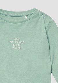 Helles grünes Langarmshirt aus weicher Stoffqualität mit rundem Halsausschnitt und zwei Druckknöpfen. Der weiße Schriftzug lautet: "LACHE UND DIE WELT LÄCHELT MIT DIR."