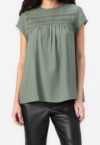 Frau in einer lockeren, kurzärmeligen olivgrünen Bluse mit Spitzendetails und schwarzen Lederhosen, die vor einem weißen Hintergrund steht.