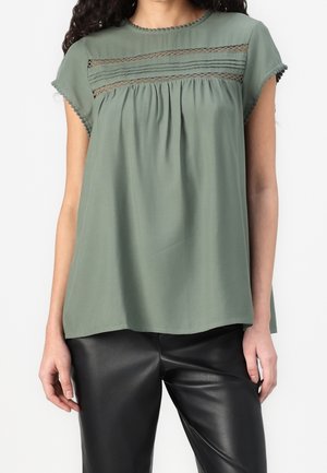 Vrouw in een losse, kortgeknipte olijvenkleurige blouse met kantdetails en zwarte leren broek, staande tegen een witte achtergrond.