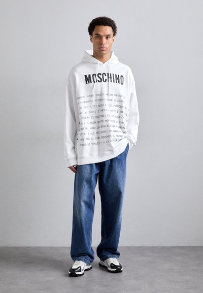 Weißer Hoodie mit schwarzem Text, ausgestattet mit einer Kapuze mit Kordelzug. Kombiniert mit lockeren blauen Jeans und schwarz-weißen Sneakers. Minimalistischer Hintergrund.