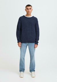 Suéter de punto azul marino con cuello redondo, corte relajado y puños acanalados, combinado con vaqueros de pierna recta azul claro y zapatillas blancas.