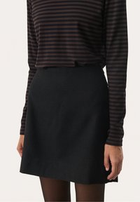 Femme portant une jupe noire trapèze, des collants noirs et un haut à manches longues rayé marron et noir, les mains détendues le long du corps.