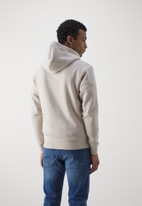 Bež hoodie izrađen od mekog materijala s kapuljačom na vezanje, rebrastim manžetama i donjim rubom. Nosivši s plavim trapericama, prikazan s leđa.