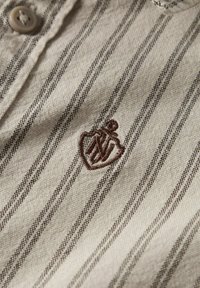 Bežasti prugasti materijal s smešnim vertikalnim linijama; sadrži dugmad za zatvaranje i smešnom smeškom izvezeni smeđi logotip u obliku srca.