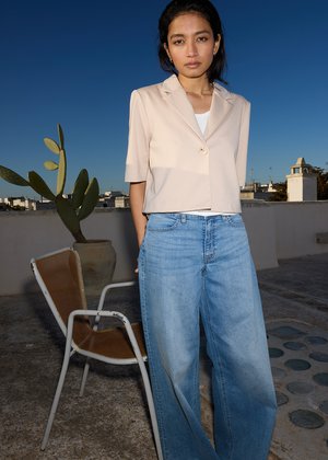 Mujer con blazer beige de manga corta y vaqueros azules de pierna ancha de pie en la azotea cerca de una silla de metal y un cactus en maceta bajo un cielo azul despejado.