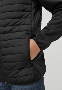 Black jacket featuring quilted texture on the upper half, with a smooth lower section and side pockets. Zipper detail on the pocket visible.