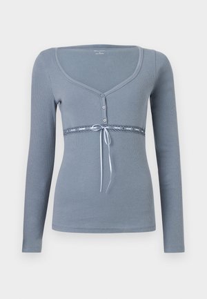 Long-sleeve, light gray top with a scoop neckline, button front, and lace detail at the waist, accented with a blue ribbon tie.