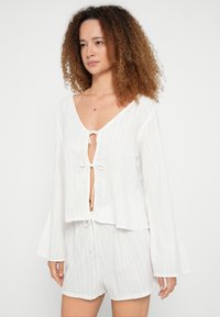 Hvid, struktureret bomulds crop top med lange klokkeærmer og knyte-lukning foran, kombineret med matchende shorts i lignende design og materiale.