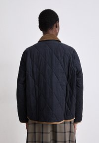 Casaco acolchoado azul-marinho com colarinho de tecido de veludo cotelê, apresentando um padrão de costura em diamante e um acabamento em tom de bege contrastante ao longo da barra e dos punhos.