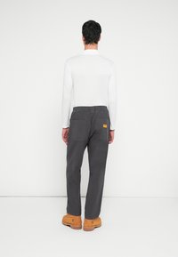 Pantaloni gri din bumbac cu talie elastică, două buzunare în spate și o mică insignă portocalie cu logo, asortați cu o bluză albă cu mâneci lungi și bocanci maro.