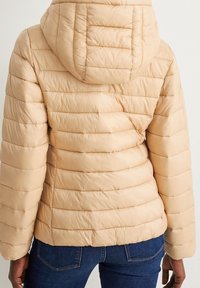 Béžová prošívaná péřová bunda s kapucí, kterou nosí osoba v modrých džínách, viděná zezadu na jednobarevném pozadí.